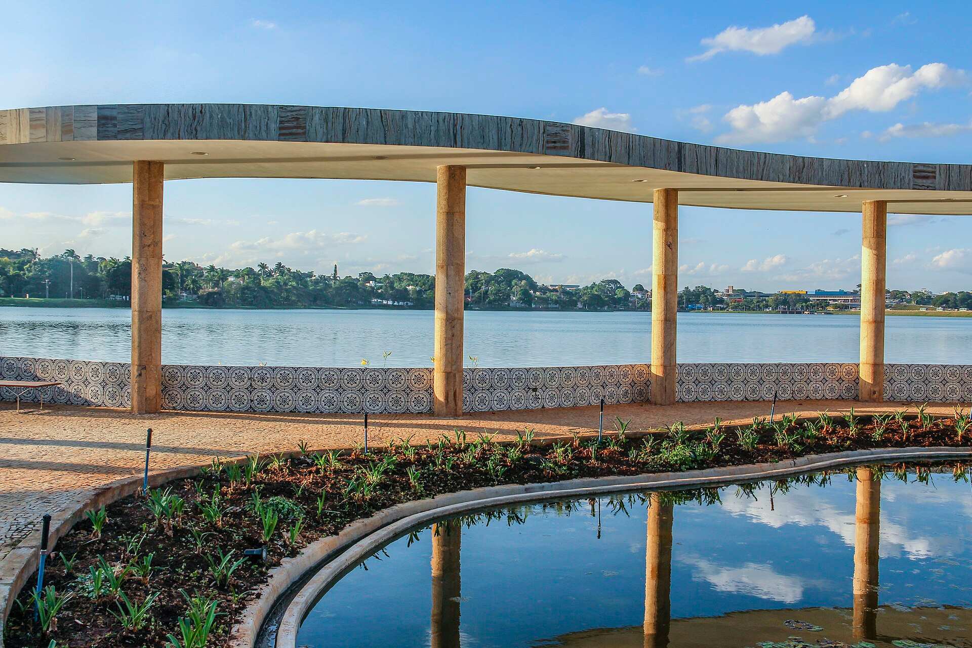 Marquise curva de colunas e muro de azulejos da Casa do Baile, com a Lagoa da Pampulha ao fundo e céu azul