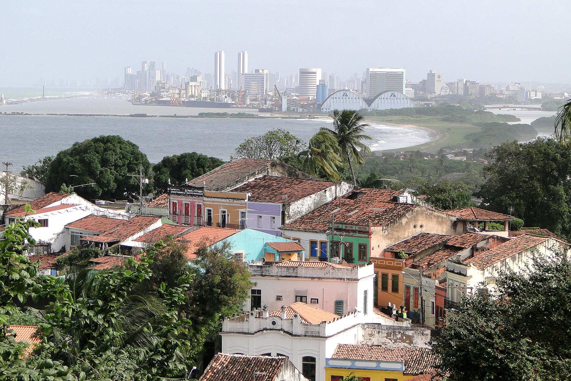 Telhados e casarões coloridos de Olinda vistos do Alto da Sé, com o mar e a silhueta de prédios do Recife ao fundo